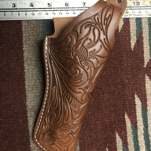 Op de afbeelding: Een bruine leren holster met ingewikkelde florale tooling en witte stiksels. De holster heeft een riemlus en een flap die de opening bedekt.