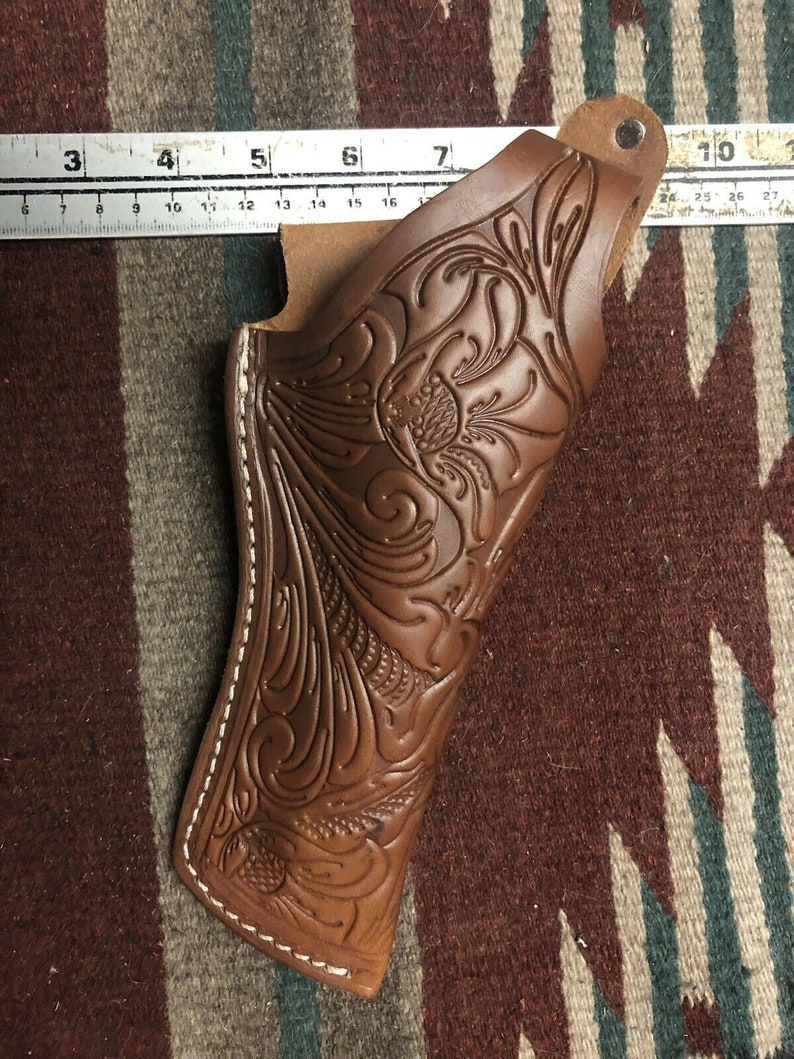 Op de afbeelding: Een bruine leren holster met ingewikkelde florale tooling en witte stiksels. De holster heeft een riemlus en een flap die de opening bedekt.