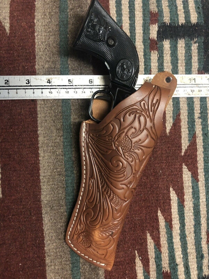 Op de afbeelding: Een bruine leren holster met ingewikkelde bloemmotieven, ontworpen om een ​​zwarte revolver te houden. De holster heeft witte stiksels rond de randen.