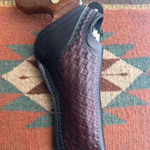 May include: A black and burgundy leather holster with a basketweave pattern, holding a revolver with a wooden grip. The holster has a black border and is set against a patterned textile background.