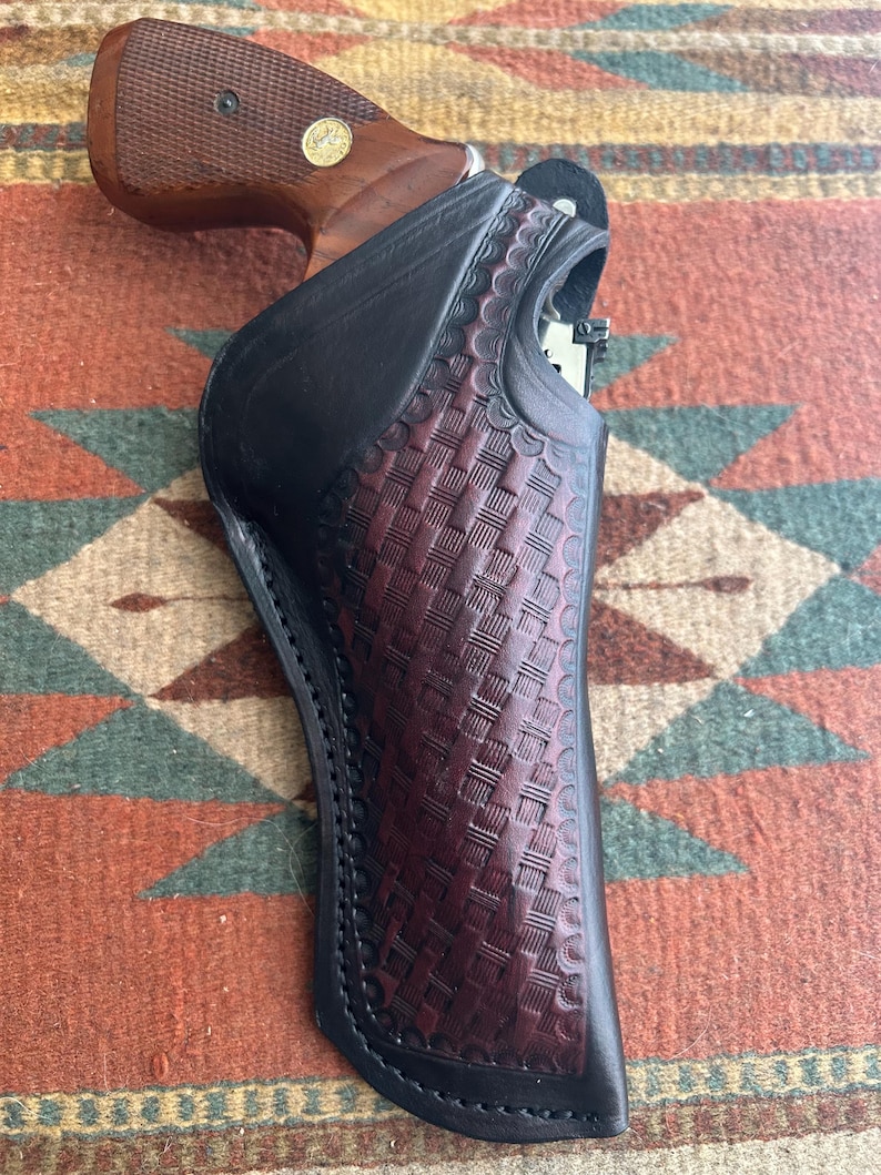 May include: A black and burgundy leather holster with a basketweave pattern, holding a revolver with a wooden grip. The holster has a black border and is set against a patterned textile background.