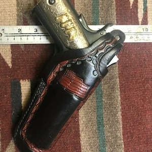 以下が含まれることがあります： 複雑な工具と茶色の革のアメリカ国旗のデザインが施された黒い革製のホルスター。ホルスターには、彫刻が施された銀色の拳銃が一部見えています。