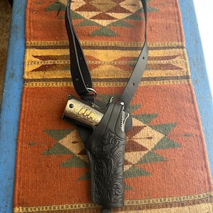 May include: A black leather shoulder holster with a decorative floral pattern, holding a silver and gold handgun. The holster is connected to a black strap. The background is a colorful patterned rug.