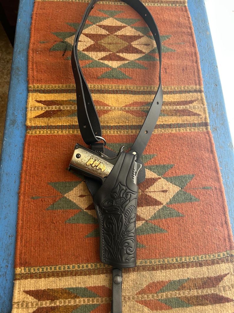 May include: A black leather shoulder holster with a decorative floral pattern, holding a silver and gold handgun. The holster is connected to a black strap. The background is a colorful patterned rug.