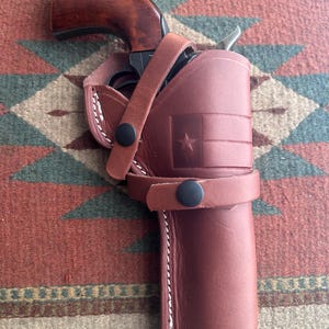 May include: A brown leather holster containing a revolver with a polished wooden grip. The holster features a stamped Texas flag design, white contrast stitching, and two black snap buttons securing the firearm in place within the leather casing.