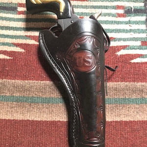 Puede incluir: Una funda de cuero negra con herramientas intrincadas y un "US" marrón en relieve en la parte delantera. Un revólver negro y dorado está en la funda.