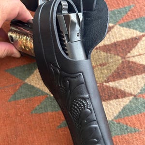 May include: Black leather holster with floral tooling, holding a silver and gold handgun. The holster has a belt loop and a strap. The handgun is partially visible, showing the barrel and grip. The background is a patterned rug.