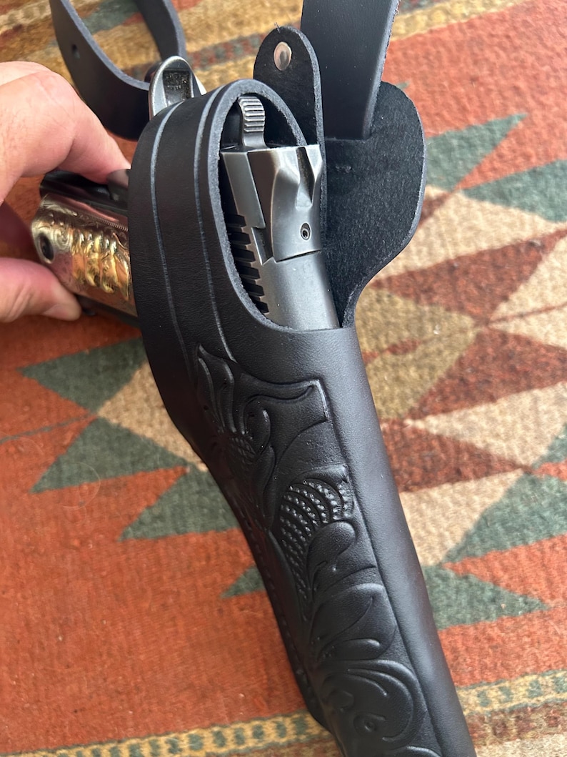 May include: Black leather holster with floral tooling, holding a silver and gold handgun. The holster has a belt loop and a strap. The handgun is partially visible, showing the barrel and grip. The background is a patterned rug.