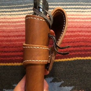 May include: A brown leather holster with a black gun inside. The holster has a flap that closes with a strap and buckle. The holster is held in a person's hand.