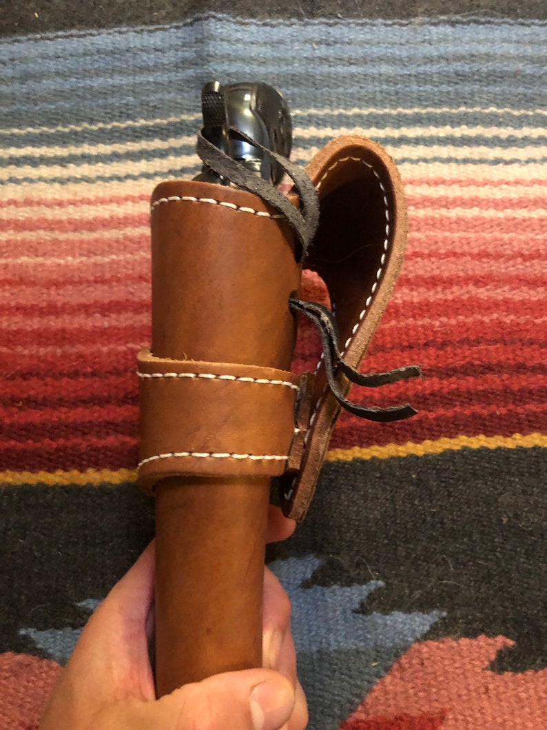 May include: A brown leather holster with a black gun inside. The holster has a flap that closes with a strap and buckle. The holster is held in a person's hand.