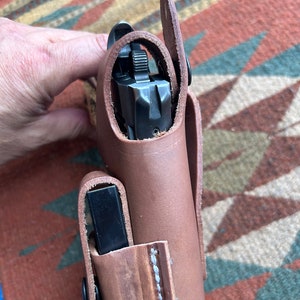 May include: A brown leather holster with a black gun inside. The holster has a flap that covers the gun and a snap closure.