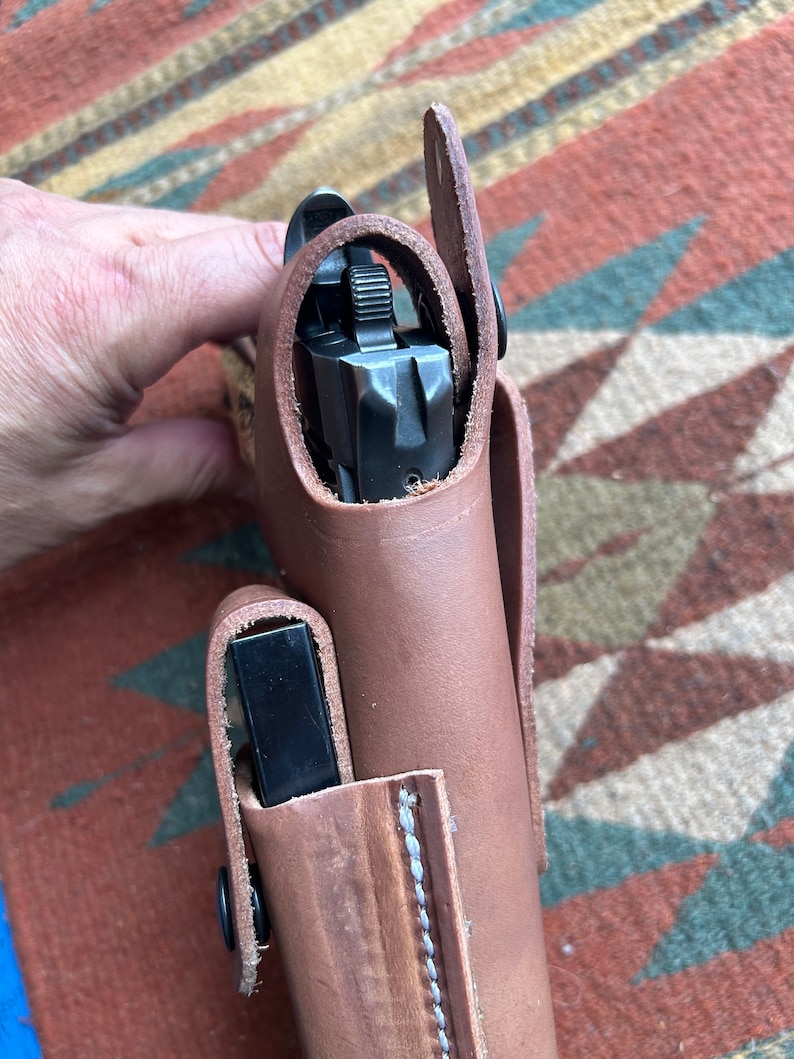 May include: A brown leather holster with a black gun inside. The holster has a flap that covers the gun and a snap closure.