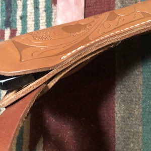 May include: A brown leather holster with intricate floral designs and stitching. The holster is folded over, revealing a black leather strap.