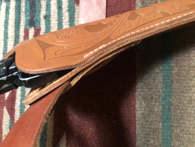 May include: A brown leather holster with intricate floral designs and stitching. The holster is folded over, revealing a black leather strap.