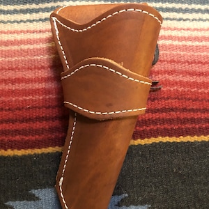 May include: A brown leather holster with white stitching. The holster is designed to hold a pistol.