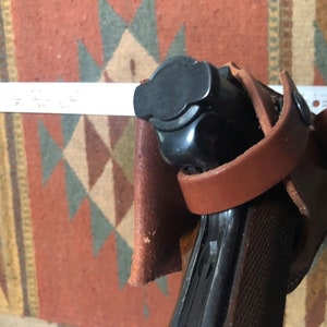 Puede incluir: Una funda de cuero marr&oacute;n con una pistola negra en su interior. La funda tiene una correa que rodea la pistola y una solapa que la cubre.