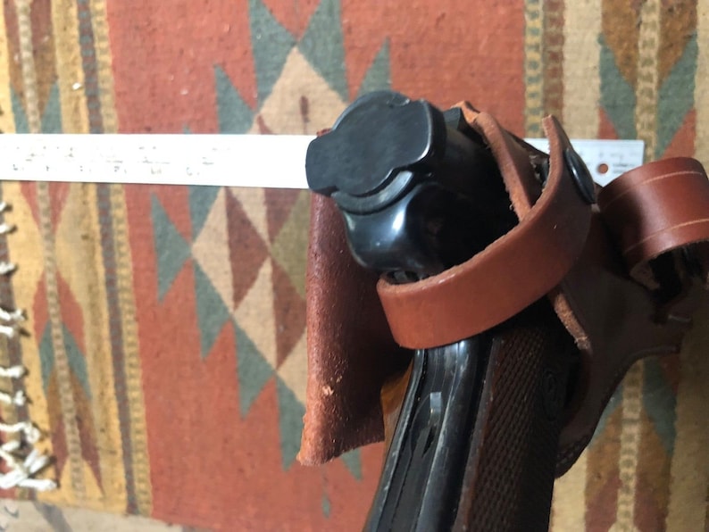 Puede incluir: Una funda de cuero marr&oacute;n con una pistola negra en su interior. La funda tiene una correa que rodea la pistola y una solapa que la cubre.