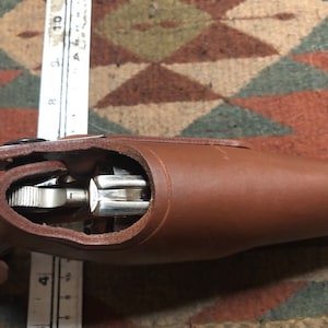 Puede incluir: Una funda de cuero marr&oacute;n con una pistola plateada en su interior. La funda est&aacute; abierta y el arma es visible.