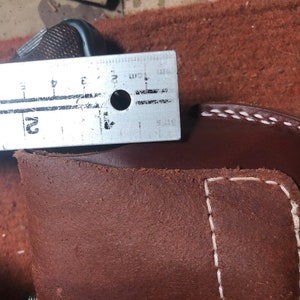 Puede incluir: Primer plano de una funda de cuero marr&oacute;n con costuras blancas. Una regla de metal se coloca encima de la funda, mostrando una medida de 5 cm.