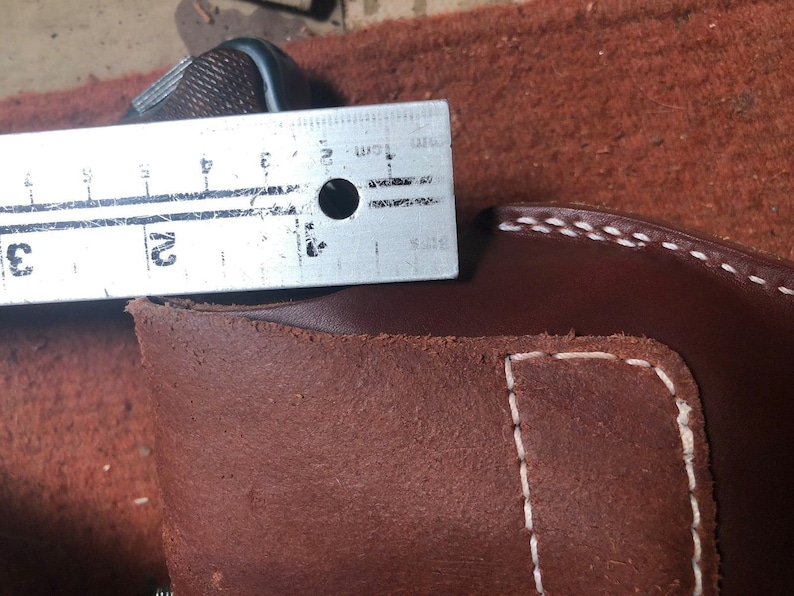 Puede incluir: Primer plano de una funda de cuero marr&oacute;n con costuras blancas. Una regla de metal se coloca encima de la funda, mostrando una medida de 5 cm.