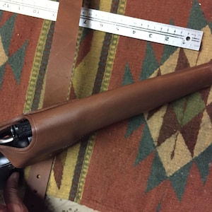 Puede incluir: Una funda de cuero marr&oacute;n con una pistola negra en su interior. La funda est&aacute; sobre una alfombra con estampado.