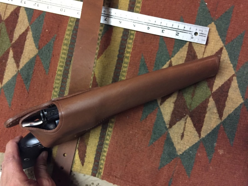 Puede incluir: Una funda de cuero marr&oacute;n con una pistola negra en su interior. La funda est&aacute; sobre una alfombra con estampado.