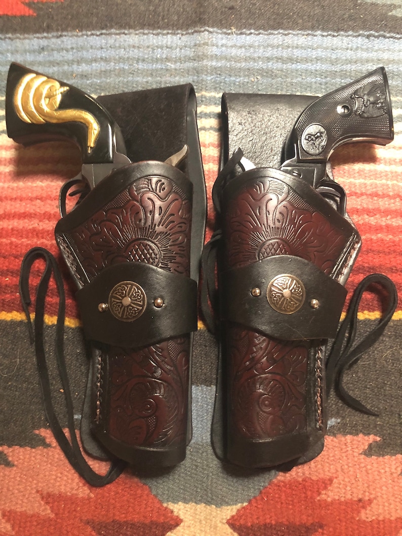 May include: Two brown leather holsters with intricate floral designs and silver accents. Each holster has a black strap and a black leather backing. The holsters are empty.