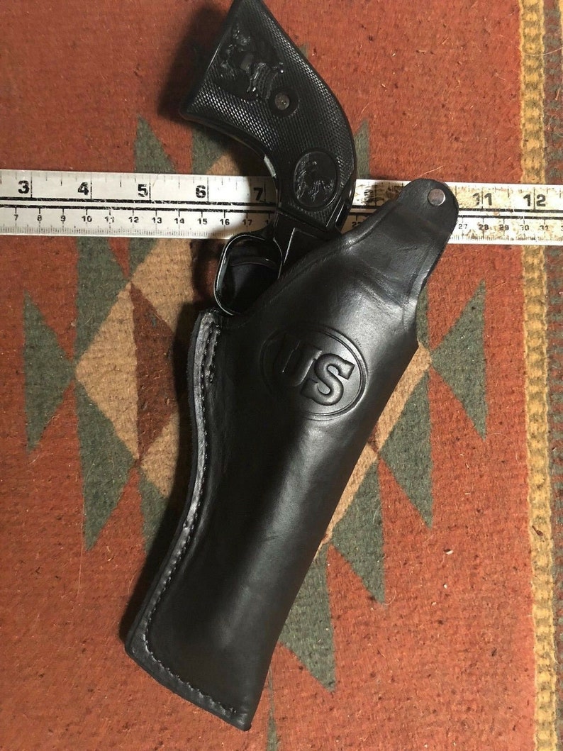 Pode incluir: Um coldre de couro preto com uma borda costurada e um bot&atilde;o de metal. O coldre tem um logotipo dos EUA em relevo na frente. Uma arma de fogo preta &eacute; parcialmente vis&iacute;vel dentro do coldre.