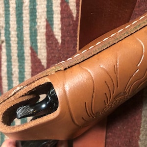 May include: A brown leather holster with a floral design and white stitching. A black handgun is partially visible inside the holster.