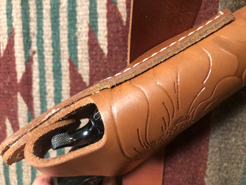May include: A brown leather holster with a floral design and white stitching. A black handgun is partially visible inside the holster.