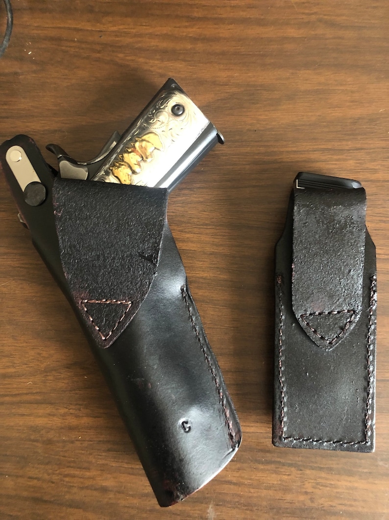 Puede incluir: Dos fundas de cuero negro para una pistola. Una funda est&aacute; abierta y tiene una pistola plateada con grabados dorados en su interior. La otra funda est&aacute; cerrada.