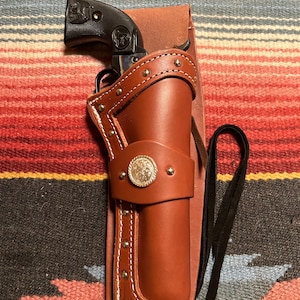 May include: A brown leather holster with a silver concho and a black pistol inside. The holster has a strap and is decorated with silver studs.