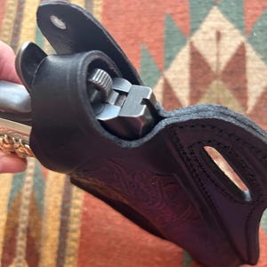 K&ouml;nnte beinhalten: Ein schwarzes Lederholster mit aufwendigen Blumenmustern. Das Holster ist f&uuml;r eine Pistole ausgelegt und hat eine Klappe, die die Waffe sichert.