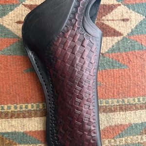 May include: A black leather holster with a burgundy basketweave pattern. The holster has a curved shape and a loop at the top. The stitching is visible along the edges. The holster is designed to hold a firearm.