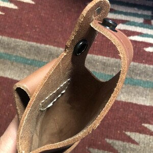 Op de afbeelding: Een bruine leren holster met een gestikte naad en drie metalen drukknopen. De holster is open en leeg.