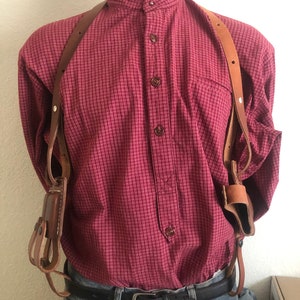 Puede incluir: Una funda de hombro de cuero marrón con dos bolsas se muestra sobre una camisa roja a cuadros con botones. La camisa está metida en unos vaqueros azules y se ve un cinturón negro. El maniquí tiene un tono de piel neutro.