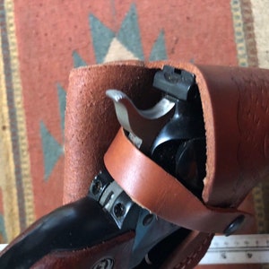 Op de afbeelding: Een bruine leren holster met een zwart revolver erin. De holster heeft een band die om de taille gaat.
