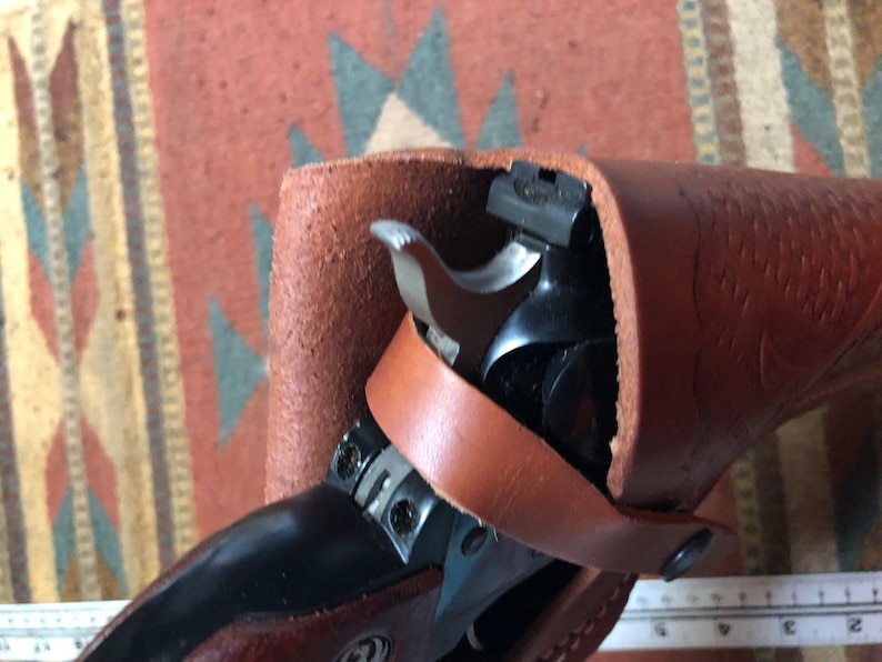 Op de afbeelding: Een bruine leren holster met een zwart revolver erin. De holster heeft een band die om de taille gaat.