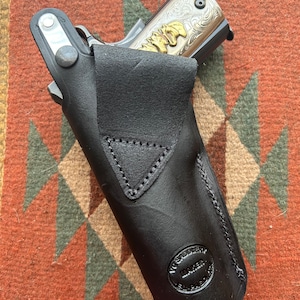 May include: A black leather holster with a silver snap and a "V. Saddlery Maker El Paso" stamp. A silver and gold engraved handgun is partially visible inside the holster.