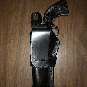 May include: A black leather holster with a black handgun inside. The holster has a flap that covers the gun and a belt loop.