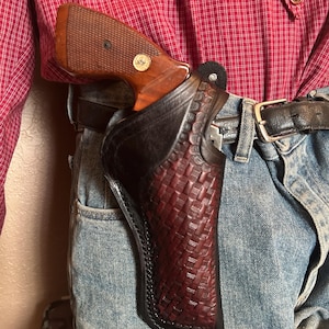 May include: A brown and black leather holster with a woven pattern, holding a handgun with a wooden grip. The holster is attached to a black belt worn with blue jeans and a red plaid shirt.