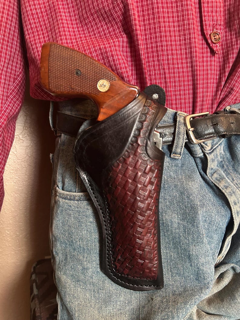 May include: A brown and black leather holster with a woven pattern, holding a handgun with a wooden grip. The holster is attached to a black belt worn with blue jeans and a red plaid shirt.