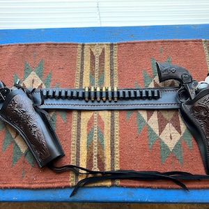 Puede incluir: Un cinturón de pistola de cuero marrón con dos fundas y un cinturón de cartuchos. El cinturón está decorado con intrincados diseños florales. Las fundas están vacías y el cinturón de cartuchos está lleno de balas.