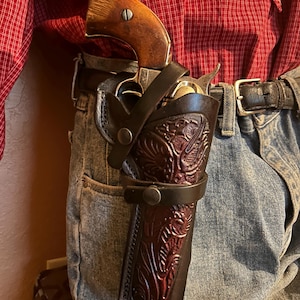 May include: A vintage revolver in a dark brown leather holster with an embossed floral design. The holster is secured to a belt, and the revolver's wooden handle is visible. The person is wearing a red plaid shirt and blue jeans.