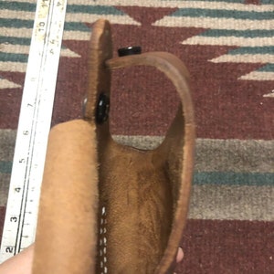 Op de afbeelding: Een bruine leren holster met een gestikte naad en een drukknopsluiting. De holster is open en leeg.