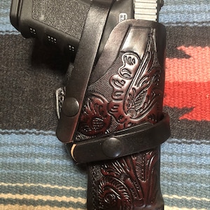 Puede incluir: Una funda de cuero marrón con intrincados detalles florales, diseñada para sostener una pistola negra. La funda tiene una correa con cierre de presión y un clip de metal.
