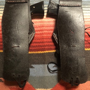 May include: Two black leather holsters with a black gun in each. The holsters have a strap and a tie at the bottom.
