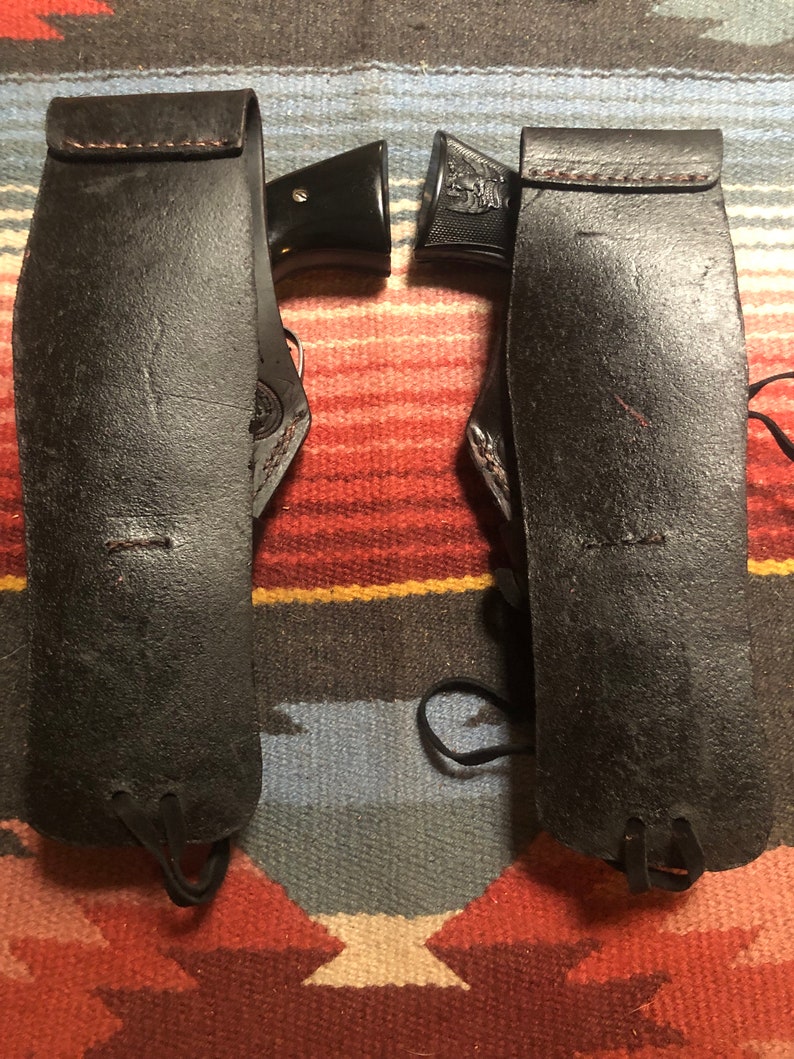 May include: Two black leather holsters with a black gun in each. The holsters have a strap and a tie at the bottom.