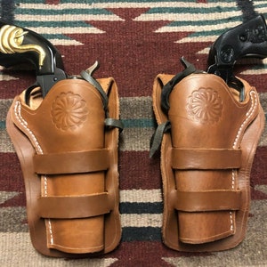 May include: Two black revolvers with gold detailing, each in a brown leather holster. The holsters have a floral pattern and are secured with straps. The background is a patterned rug.