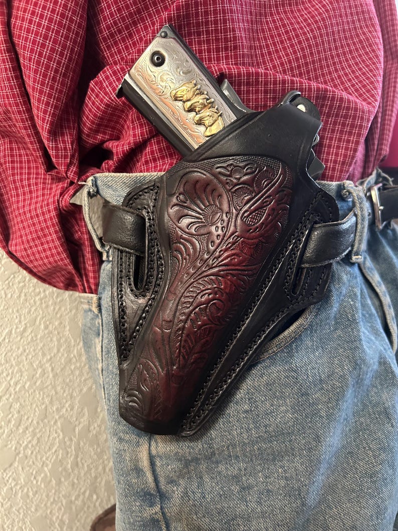 K&ouml;nnte beinhalten: Ein schwarzes Lederholster mit aufwendigen floralen Verzierungen, entworfen, um eine silberne Pistole mit goldenen Akzenten zu halten. Das Holster wird an einem G&uuml;rtel getragen.